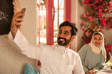 Taking commemorative selfies using a mobile phone, a dining table filled with a variety of delicious dishes and appetizers, gestures of happiness while spending enjoyable times with friends, a gathering of friends at a delicious breakfast table, a close-up picture of a lunch invitation for Saudi Gulf Arab friends enjoying food at a luxury restaurant.