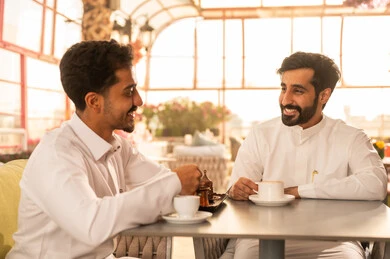 تعابير السعادة والسرور بلمة الرفاق، مشاركة المشروبات الساخنة مع الاصدقاء ، كوفي شوب راقي بالمملكة العربية السعودية، تطوير العلاقات الإجتماعية، قضاء وقت ممتع مع الأصدقاء، الاستمتاع بالاجواء السعيده ، صديقان عربيان خليجيان سعوديان يرتديان الثوب الابيض يجلسان على طاولة في المقهى