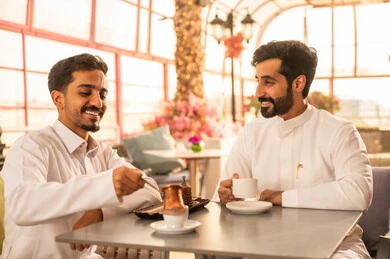 تطوير العلاقات الإجتماعية، مشاركة المشروبات الساخنة مع الاصدقاء ، كوفي شوب راقي بالمملكة العربية السعودية، تعابير السعادة والسرور بلمة الرفاق،قضاء وقت ممتع مع الأصدقاء، الاستمتاع بالاجواء السعيده ، صديقان عربيان خليجيان سعوديان يرتديان الثوب الابيض يجلسان على طاولة في المقهى