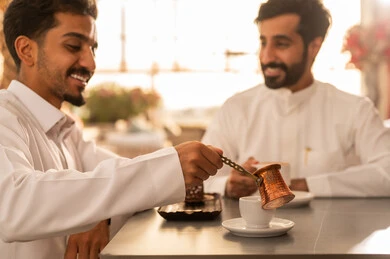 كوفي شوب راقي بالمملكة العربية السعودية، مشاركة المشروبات الساخنة مع الاصدقاء ،تعابير السعادة والسرور بلمة الرفاق، تطوير العلاقات الإجتماعية، قضاء وقت ممتع مع الأصدقاء، الاستمتاع بالاجواء السعيده ، صديقان عربيان خليجيان سعوديان يرتديان الثوب الابيض يجلسان على طاولة في المقهى