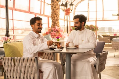 Sharing hot drinks with friends, a classy coffee shop in the Kingdom of Saudi Arabia, expressions of happiness and joy in the company of companions, developing social relationships, spending enjoyable time with friends, enjoying the happy atmosphere, two Arab Gulf friends from Saudi Arabia wearing white thobes sitting at a table in the café.