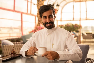 Spending free time sipping a favorite drink, enjoying warm winter beverages, relishing hot drinks, an Arab Gulf Saudi man wearing a white thobe sitting in a classy coffee shop drinking coffee, gestures of happiness and joy.