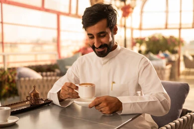 تناول المشروبات الشتوية الدافئة، قضاء وقت الفراغ باحتساء المشروب المفضل،الاستمتاع بالمشاريب الساخنة، رجل عربي خليجي سعودي يرتدي الثوب الأبيض يجلس في كوفي شوب راقي يقوم بشرب القهوة، إيماءات السعادة والسرور