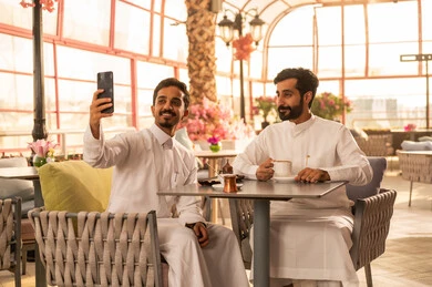 Documenting happy moments, taking commemorative selfies using a mobile phone, sharing hot drinks with friends, an upscale coffee shop in the Kingdom of Saudi Arabia, expressions of happiness and joy with friends, developing social relationships, spending enjoyable time with friends, enjoying the happy atmosphere, two Arab Gulf friends from Saudi Arabia wearing white thobes sitting at a table in the café.