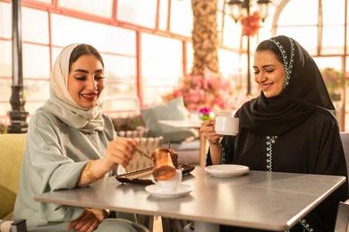 قضاء وقت ممتع مع الأصدقاء، مشاركة المشروبات الساخنة مع الاصدقاء ، كوفي شوب راقي بالمملكة العربية السعودية، تعابير السعادة والسرور بلمة الرفاق، تطوير العلاقات الإجتماعية، الاستمتاع بالاجواء السعيده ، صديقتان عربيتان خليجيتان سعوديتان تجلسان على طاولة في المقهى