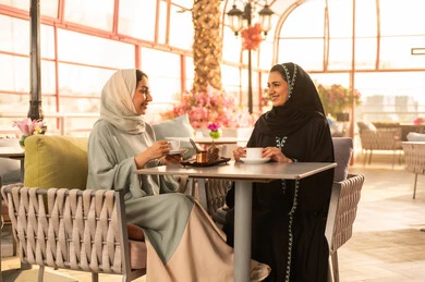 مشاركة المشروبات الساخنة مع الاصدقاء ، كوفي شوب راقي بالمملكة العربية السعودية، تعابير السعادة والسرور بلمة الرفاق، تطوير العلاقات الإجتماعية، قضاء وقت ممتع مع الأصدقاء، الاستمتاع بالاجواء السعيده ، صديقتان عربيتان خليجيتان سعوديتان تجلسان على طاولة في المقهى