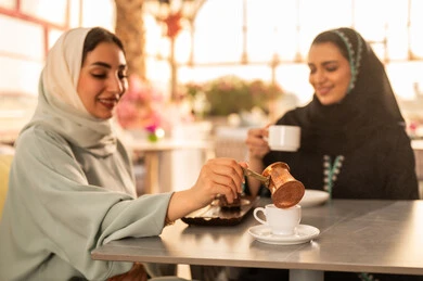 صديقتان عربيتان خليجيتان سعوديتان تجلسان على طاولة في المقهى، مشاركة المشروبات الساخنة مع الاصدقاء ، كوفي شوب راقي بالمملكة العربية السعودية، تعابير السعادة والسرور بلمة الرفاق، تطوير العلاقات الإجتماعية، قضاء وقت ممتع مع الأصدقاء، الاستمتاع بالاجواء السعيده