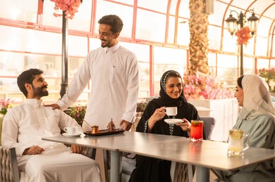 A group of Arab Gulf friends from Saudi Arabia are sitting at a table in a café, a high-end coffee shop in the Kingdom of Saudi Arabia. Expressions of happiness and joy among the companions, developing social relationships, spending enjoyable time with friends, enjoying hot drinks, and enjoying the happy atmosphere.