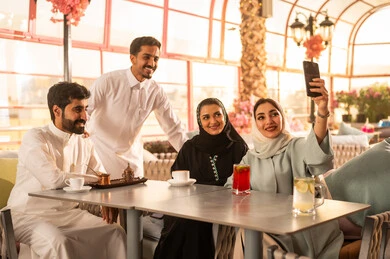 التقاط صور تذكارية بواسطة الجوال ، توثيق اللحظات السعيدة ، كوفي شوب راقي بالمملكة العربية السعودية، تعابير السعادة والسرور بلمة الرفاق، تطوير العلاقات الإجتماعية، قضاء وقت ممتع مع الأصدقاء، الاستمتاع بالمشروبات الساخنه ، الاستمتاع بالاجواء السعيده ، مجموعة اصدقاء عرب خليجيين سعوديين يجلسون على طاولة في المقهى