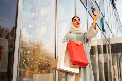 The use of modern technologies, gestures of joy and happiness, pride and appreciation for architectural progress, urban growth and development in Saudi Arabia, strolling and breathing fresh air outside, an Arab Gulf Saudi woman wearing an abaya and hijab holding a mobile phone and taking a selfie.