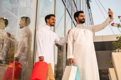 Gestures of joy and happiness, strolling and breathing in the fresh air outside, pride and appreciation for architectural progress, growth and urban development in Saudi Arabia, two Arab Gulf Saudi men wearing white thobes taking a commemorative photo using a mobile phone.