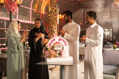 A Saudi Gulf Arab family is celebrating their friend's birthday at a café, with a joyful and festive atmosphere, participating in birthday celebrations, a decorated cake with candles, the concept of enjoyment and entertainment, expressions of affection and warmth, celebrating birthdays.