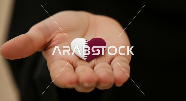 The concept of love and belonging to the country, national holidays and occasions, commemorating the founding of the state, celebrating Qatar National Day on December 18, a close-up image of a Qatari Gulf Arab woman wearing a hijab holding the Qatari flag shaped like a heart in her hand with gestures of pride and belonging, national symbols and cultures, love for the homeland and belonging.