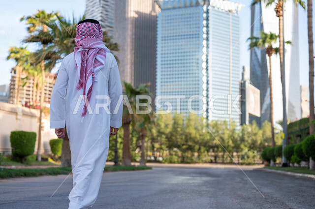 صورة من الخلف لشاب عربي خليجي سعودي يرتدي الزي التقليدي يقوم بجولة داخل مدينة الملك عبدالله المالية ( كافد ) في مدينة الرياض في المملكة العربية السعوية ، إطلالة رائعة لأبراج المدينة ، جولة سياحية في المدينة