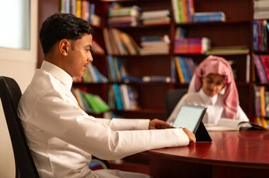 The use of tablets in education, education and teaching in the Kingdom of Saudi Arabia, the importance of knowledge in developing mental abilities, the concept of diligence and the pursuit of excellence, gestures of integration and focus, two Arab Gulf Saudi students wearing white thobes sitting inside the school library.