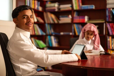 Education in the Kingdom of Saudi Arabia, the importance of knowledge in developing mental abilities, the concept of diligence and the pursuit of excellence, gestures of integration and focus, two Arab Gulf Saudi students wearing white thobes sitting inside the school library, using tablets in studying.
