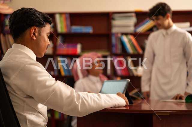استخدام جهاز التابلت، اداء الواجبات المدرسية، الاستفادة من التقنيات والتكنولوجيا الحديثة في التعلم والدراسة، المناهج المطورة والتعليم الالكتروني، تطور طرق التعليم ، حب التعلم واستكشاف الأفكار، مجموعه من الطلاب العرب الخليجيون السعوديون يرتدون الثوب الابيض يجلسون داخل المكتبة المدرسية برفقه معلمهم