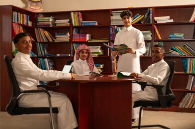 Studying and discussing lessons, teamwork and cooperation among colleagues, using tablets in education, the concept of diligence and striving for excellence, education in the Kingdom of Saudi Arabia, a group of Arab Gulf students from Saudi Arabia wearing white thobes sitting inside the school library.