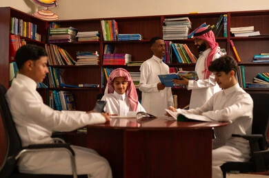 الاستفادة من الأدوات التكنولوجية في التعلم، المناهج المطورة والتعليم الالكتروني، تطور طرق التعليم ، حب التعلم واستكشاف الأفكار، مجموعه من الطلاب العرب الخليجيون السعوديون يرتدون الثوب الابيض يجلسون داخل المكتبة المدرسية برفقه معلمهم، استخدام جهاز التابلت، اداء الواجبات المدرسية