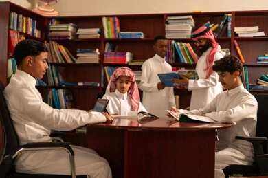 Using technology to support learning and studying, developed curricula and e-learning, the evolution of teaching methods, a love for learning and exploring ideas, a group of Arab Gulf Saudi students wearing white thobes sitting inside the school library with their teacher, using a tablet, doing homework.
