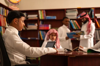 The use of modern technologies in academic education, developed curricula and e-learning, the evolution of teaching methods, the love of learning and exploring ideas, a group of Arab Gulf Saudi students wearing white thobes sitting inside the school library with their teacher, using a tablet, doing homework.