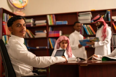 الاستفادة من التكنولوجيا في العملية التعليمية، المناهج المطورة والتعليم الالكتروني، تطور طرق التعليم ، حب التعلم واستكشاف الأفكار، مجموعه من الطلاب العرب الخليجيون السعوديون يرتدون الثوب الابيض يجلسون داخل المكتبة المدرسية برفقه معلمهم، استخدام جهاز التابلت، اداء الواجبات المدرسية