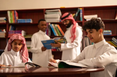 طلاب عربيين سعوديين خليجيين يرتدون الثوب الابيض يجلسون في المكتبة المدرسية برفقة معلمهم، معلم عربي خليجي سعودي يقوم بمساعده طلابه، الاهتمام بالقراءة المدرسية، التعليم في المملكة العربية السعودية،  تأدية الواجبات المدرسية، الحوارات الودية بين المعلم والطالب