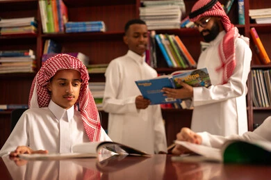 التعليم في المملكة العربية السعودية،  تأدية الواجبات المدرسية، الاستمتاع بقراءة الكتب المدرسية، الحوارات الودية بين المعلم والطالب ، طلاب عربيين سعوديين خليجيين يرتدون الثوب الابيض يجلسون في المكتبة المدرسية برفقة معلمهم ، معلم عربي خليجي سعودي يقوم بمساعده طلابه