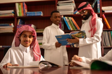 تأدية الواجبات المدرسية، الاستمتاع بقراءة الكتب المدرسية، الحوارات الودية بين المعلم والطالب ، طلاب عربيين سعوديين خليجيين يرتدون الثوب الابيض يجلسون في المكتبة المدرسية برفقة معلمهم ، معلم عربي خليجي سعودي يقوم بمساعده طلابه، التعليم في المملكة العربية السعودية
