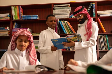 Enjoying reading school books, friendly dialogues between the teacher and the student, two Saudi Arabian Gulf students wearing white thobes sitting in the school library with their teacher, doing homework, a Saudi Arabian Gulf teacher helping his students, education in the Kingdom of Saudi Arabia.
