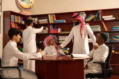 Reliance on modern technologies in learning, developed curricula and e-learning, the evolution of teaching methods, love of learning and exploring ideas, a group of Arab Gulf Saudi students wearing white thobes sitting inside the school library with their teacher, using a tablet, doing homework.