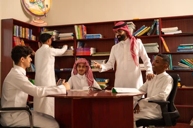 توظيف التكنولوجيا الحديثة في الدراسة، المناهج المطورة والتعليم الالكتروني، تطور طرق التعليم ، حب التعلم واستكشاف الأفكار، مجموعه من الطلاب العرب الخليجيون السعوديون يرتدون الثوب الابيض يجلسون داخل المكتبة المدرسية برفقه معلمهم، استخدام جهاز التابلت، اداء الواجبات المدرسية