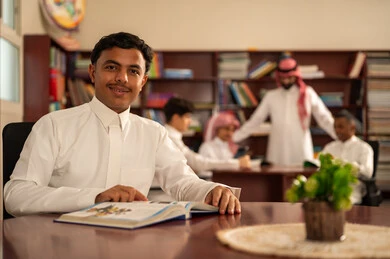 The use of technological means in learning, developed curricula and e-learning, the evolution of teaching methods, the love of learning and exploring ideas, a group of Arab Gulf Saudi students wearing white thobes sitting inside the school library with their teacher, using a tablet, doing homework.
