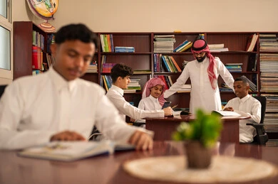 The use of digital technologies in education, developed curricula and e-learning, the evolution of teaching methods, the love of learning and exploring ideas, a group of Arab Gulf Saudi students wearing white thobes sitting inside the school library with their teacher, using a tablet, doing homework.