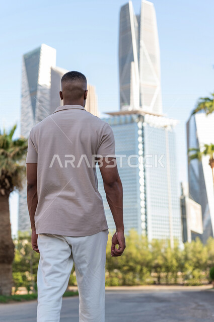 صورة من الخلف لشاب عربي خليجي سعودي ذو بشرة سمراء يقوم بجولة داخل مدينة الملك عبدالله المالية ( كافد ) في مدينة الرياض في المملكة العربية السعوية ، إطلالة رائعة لأبراج المدينة ، جولة سياحية في المدينة