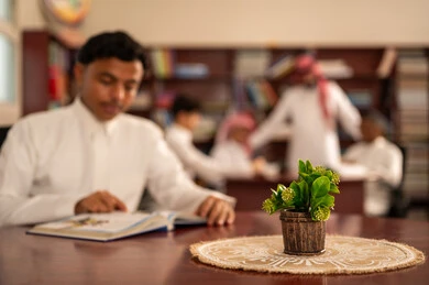 Utilizing technological means in education, developed curricula and e-learning, the evolution of teaching methods, a love for learning and exploring ideas, a group of Arab Gulf Saudi students wearing white thobes sitting inside the school library with their teacher, using a tablet, completing homework assignments.