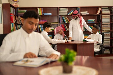 دمج التكنولوجيا في عملية التعلم، المناهج المطورة والتعليم الالكتروني، تطور طرق التعليم ، حب التعلم واستكشاف الأفكار، مجموعه من الطلاب العرب الخليجيون السعوديون يرتدون الثوب الابيض يجلسون داخل المكتبة المدرسية برفقه معلمهم، استخدام جهاز التابلت، اداء الواجبات المدرسية