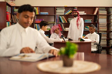 The use of modern technologies in education, developed curricula, and e-learning, the evolution of teaching methods, a love for learning and exploring ideas, a group of Arab Gulf Saudi students wearing white thobes sitting inside the school library with their teacher, using a tablet, completing homework assignments.