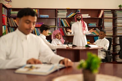 استخدام التكنولوجيا الحديثة في التعلم، المناهج المطورة والتعليم الالكتروني، تطور طرق التعليم ، حب التعلم واستكشاف الأفكار، مجموعه من الطلاب العرب الخليجيون السعوديون يرتدون الثوب الابيض يجلسون داخل المكتبة المدرسية برفقه معلمهم، استخدام جهاز التابلت، اداء الواجبات المدرسية