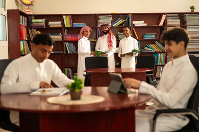 حب التعلم واستكشاف الأفكار، تطور طرق التعليم ،المناهج المطورة والتعليم الالكتروني، الاستفادة من التقنيات والتكنولوجيا الحديثة في التعلم والدراسة، مجموعه من الطلاب العرب الخليجيون السعوديون يرتدون الثوب الابيض يجلسون داخل المكتبة المدرسية برفقه معلمهم، استخدام جهاز التابلت، اداء الواجبات المدرسية