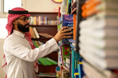 حب المطالعة والقراءة، تصفح الكتب الهادفة والمعرفية، التعليم في المملكة العربيه السعودية ، ممارسة الهوايات المفضلة ، معلم عربي خليجي سعودي يرتدي الثوب الابيض و الشماغ يقف في المكتبة المدرسية يقوم باختيار كتاب