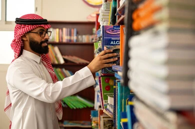 مطالعة الكتب الهادفة والمعرفية، مفهوم المطالعة والثقافة ، التعليم في المملكة العربيه السعودية ، ممارسة الهوايات المفضلة ، معلم عربي خليجي سعودي يرتدي الثوب الابيض و الشماغ يقف في المكتبة المدرسية يقوم باختيار كتاب