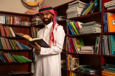 Practicing favorite hobbies, education in the Kingdom of Saudi Arabia, reading useful books, the concept of reading and culture, a Gulf Arab Saudi teacher wearing a white thobe and a ghutrah stands in the school library holding a book in his hands.