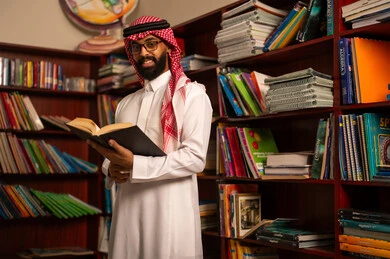 التعليم في المملكة العربية السعودية، قراءة الكتب المفيدة، مفهوم المطالعة والثقافة، ممارسة الهوايات المفضلة ، معلم عربي خليجي سعودي يرتدي الثوب الابيض و الشماغ يقف في المكتبة المدرسية يمسك بيدية كتاب