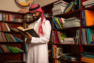 قراءة الكتب المفيدة، مفهوم المطالعة والثقافة، التعليم في المملكة العربية السعودية، ممارسة الهوايات المفضلة ، معلم عربي خليجي سعودي يرتدي الثوب الابيض و الشماغ يقف في المكتبة المدرسية يمسك بيدية كتاب