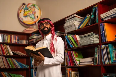 مفهوم المطالعة والثقافة ، قراءة الكتب المفيدة، التعليم في المملكة العربية السعودية، ممارسة الهوايات المفضلة ، معلم عربي خليجي سعودي يرتدي الثوب الابيض و الشماغ يقف في المكتبة المدرسية يمسك بيدية كتاب
