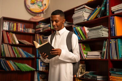 الشغف بالمعرفة والاستطلاع، زيارة مكتبة المدرسة، طالب عربي خليجي سعودي يرتدي الثوب الابيض و الحقيبة المرسية يقف امام مكتبة خشبية يحمل بيده كتاب، تنمية مهارة القراءة للطلاب، التعليم والدراسة في السعودية ، قضاء وقت الفراغ بالتعلم والتثقف الذاتي