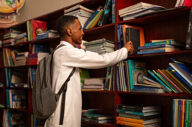 التعليم والدراسة في المملكة العربيه السعودية، تنمية مهارة المطالعة للطلاب، قضاء وقت الفراغ بالتعلم والتثقف الذاتي، الاهتمام بتصفح الكتب المفيدة, حب الثقافة والاستطلاع، طالب عربي سعودي خليجي يرتدي الثوب الابيض و الحقيبة المدرسية يقوم باختيار كتاب من المكتبة الخشبية, زيارة مكتبة المدرسة