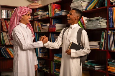 Welcoming and greeting, supporting friendly relationships among students, preparing for the new school semester, play and recreation time, two Arab Gulf Saudi students wearing white thobes and school bags standing in the school library shaking hands, gestures of happiness and fun, academic schools in Saudi Arabia.