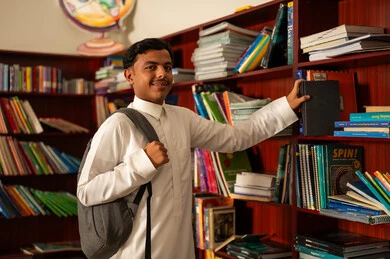 The interest in browsing useful books, developing reading skills for students, love for culture and exploration, an Arab Saudi Gulf student wearing a white thobe and a school bag is choosing a book from the wooden library, spending free time learning and self-education, visiting the school library, education and study in the Kingdom of Saudi Arabia.
